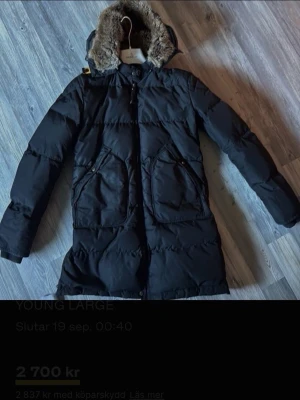 Svart lång dunjacka med pälshuva - Säljer en svart lång dunjacka från Moncler med stor huva kantad av fuskpäls. Jackan har två stora fickor framtill, dragkedja och tryckknappar samt en patch på ena ärmen. Perfekt för kalla vinterdagar och riktigt snygg streetstil.