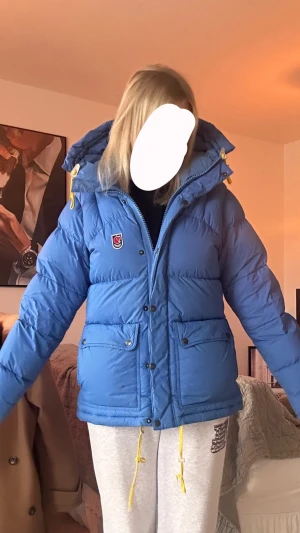 Blå dunjacka från Fjällräven - Blå dunjacka från Fjällräven med huva och gula detaljer på snören och dragkedjor. Jackan har två stora framfickor med knappar, broderad Fjällräven-logga på bröstet och en puffig, varm design perfekt för kalla dagar.