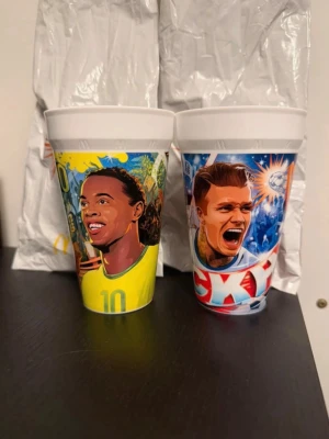 McDonald’s mugg Ronaldinho och Beckham - Säljer två färgglada plastmuggar med tryck av tecknade fotbollsspelare. Ena muggen har gul bakgrund och motiv av en spelare i gul tröja med nummer 10, den andra har blå bakgrund och en spelare som skriker med fotbollstema. Perfekta för sportfans!