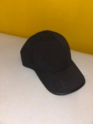 Svart keps med Fendi-mönster - Snygg svart keps från Fendi med det klassiska FF-mönstret över hela kepsen. Kepsen har böjd skärm och är tillverkad i ett tygmaterial som ger en exklusiv känsla. Perfekt accessoar för dig som vill sticka ut med stil.