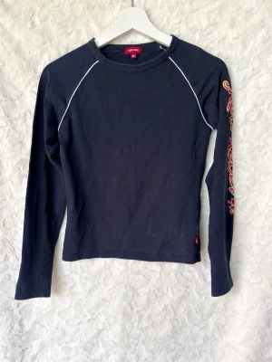 marinblå långärmad topp med tryck - Marinblå långärmad topp med vita sömdetaljer och färgglatt tryck längs ena ärmen. Modellen har en rund halsringning och är tillverkad i mjukt bomullsmaterial. Perfekt för dig som gillar en sportig och edgy stil.