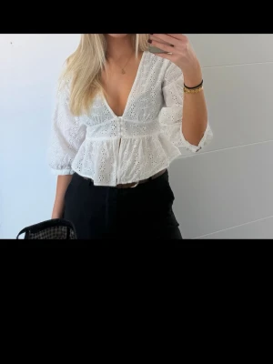 Vit broderad blus med v-ringning - Söt vit blus med broderade hålmönster och djup v-ringning. Blusen har trekvartsärm och volang nedtill Perfekt att styla med både jeans och kjol.