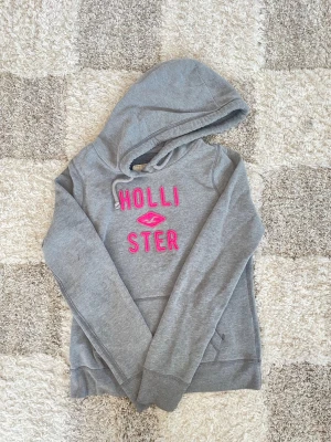 Grå hoodie från Hollister - Snygg grå hoodie från Hollister med huva och dragsko. Framtill står det 'HOLLISTER' i upphöjd rosa text och en broderad logga i mitten. Hoodien har en klassisk känguruficka och mjukt material som känns skönt mot huden. Säljer den då den inte kom till användning pris går att sänka. Passformen passar även bra S