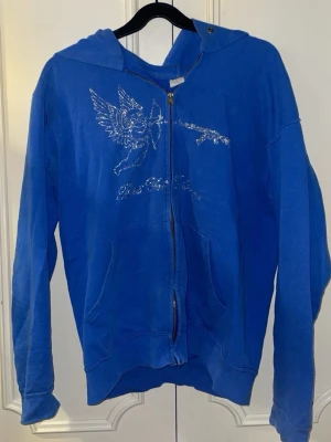 Blå hoodie med glittrigt motiv - Blå hoodie med dragkedja och huva. På bröstet finns ett glittrigt tryck av en ängel med pilbåge och text under. 