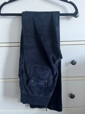 Mörkblå mjukisbyxor med ficka - Mörkblå Juicy Couture mjukisbyxor i mjukt material med breda muddar vid benslut och en ficka med lock och tryckknappar på sidan. Perfekta för chill dagar hemma eller när du vill vara bekväm. Klassisk rak passform och enkel design. Saknar lappar!