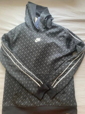 Nike  - Snygg svart hoodie från Nike med vit logga på bröstet och all-over print med små Nike-loggor. Hoodien har huva, ribbade muddar och vita detaljer längs ärmarna. Perfekt för en sportig och avslappnad stil.