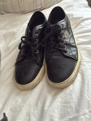 Svarta låga sneakers i läder - Säljer ett par svarta låga sneakers med ovandel i slätt läder och vita sulor. Skorna har svarta skosnören och metallfärgade öljetter. Klassisk och enkel design som passar till det mesta.
