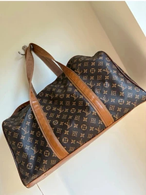 Brun monogram weekendbag - Snygg brun weekendbag med klassiskt monogrammönster och detaljer i ljusbrunt läder. Väskan har två långa handtag i tyg och läder, perfekt för resor eller gymmet. Rymlig och stilren design som sticker ut.
