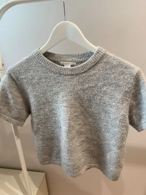 Grå stickad kortärmad tröja - Säljer en stilren grå stickad tröja med korta ärmar och rund hals. Tröjan har en mjuk och mysig känsla, perfekt för lager på lager. Ribbstickad kant vid halsen ger en snygg detalj. Passar till både jeans och kjol.