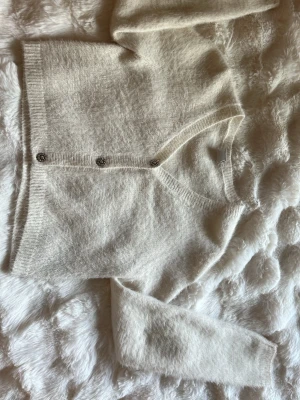 Vit fluffig kofta med dekorativa knappar - Supersöt vit kofta i fluffigt material med v-ringning och tre dekorativa silverfärgade knappar framtill. Kortare modell med ribbade muddar vid ärmslut och nederkant. Perfekt att slänga över en topp för en mysig och trendig look.