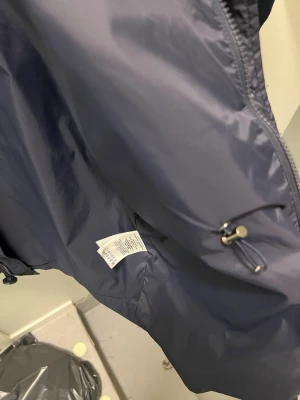 Mörkblå vindjacka från Moncler - Snygg mörkblå vindjacka från Moncler med dragkedja, tryckknappar och justerbar dragsko i midjan. Jackan har Moncler-logga på ärmen och är tillverkad i ett lätt, glansigt material som står emot vind. Perfekt för dig som vill ha en stilren och funktionell jacka.