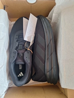 Svarta Adidas sneakers med mesh - Snygga svarta sneakers från Adidas med klassiska tre ränder på sidan. Ovandelen är i mesh och syntetmaterial för bra andningsförmåga och komfort. Tjock sula med bra grepp och dämpning. Perfekta för dig som gillar sportig stil.