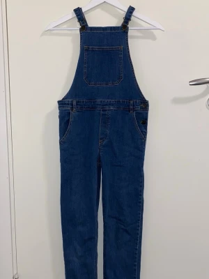 Klassisk blå jeansjumpsuit - Säljer en klassisk blå jeansjumpsuit med raka ben och justerbara hängslen. Den har en stor bröstficka framtill och sidofickor, samt knappar i metall vid midjan. Materialet är denim i 100% bomull och modellen har en avslappnad passform.