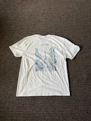 Vit t-shirt med Paris-tryck - Vit t-shirt med ett stort blått tryck på ryggen som föreställer Eiffeltornet och en parisiskt gata. Texten 'PARIS' står ovanför motivet. Klassisk passform och korta ärmar, perfekt för dig som gillar stilrena och urbana motiv.