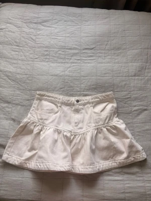 Vit jeans volangkjol med fickor - Söt vit kortkjol med volang och två framfickor. Kjolen har knapp och dragkedja framtill samt markerad söm i midjan. Kjolen ger looken av en jeanskjol men är gjord i bomull och därför väldigt skön och lätt att bära.
