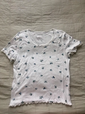 Vit blommig topp från Kappahl - Söt vit topp från Kappahl med små blå blommor och diskreta ränder. Toppen har korta ärmar, rund halsringning med en liten rosett och vågig kant nedtill. Kan användas som vanlig topp eller pyjamas.