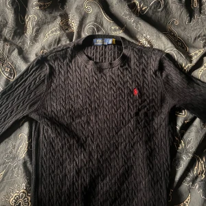  kabelstickad Polo Ralph Lauren - Kabelstickad svart tröja från Polo Ralph Lauren med röd broderad logga på bröstet. Klassisk och tidlös design med ribbade muddar vid ärmslut och nederkant. Perfekt, enkel att matcha.