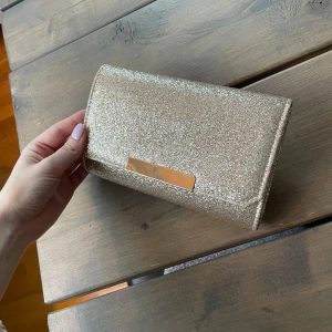 Glittrig guld kuvertväska från Mascara - Snygg glittrig kuvertväska i guld från Mascara.  Väskan har en elegant kedja och en stilren design med en metallplatta framtill. Insidan är rymlig nog för dina nödvändigheter. Längd 20cm. (Köpt på Plick och återanvänder därför bilder från den tidigare säljaren.) 