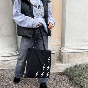 Flash Bag Black - Svart tygväska tillverkad i 100% bomull med ett tryck bestående av 6 blixtrar i en silverfärgad reflex. Centrerat på baksidan är även vår logotyp tryckt i silverfärgad reflex.