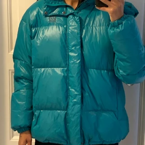 66 North puffer - Turkis dunjakke med hætte. Lynlås og knaplukning. Elastik i bunden og i hætten, som kan justeres. 2 lommer