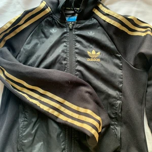 Adidas tröja med luva - Adidas tröja med guldiga sträck och luva. De är ett glansigare material fram till med adidas loggan. Använd skick. Storlek 36