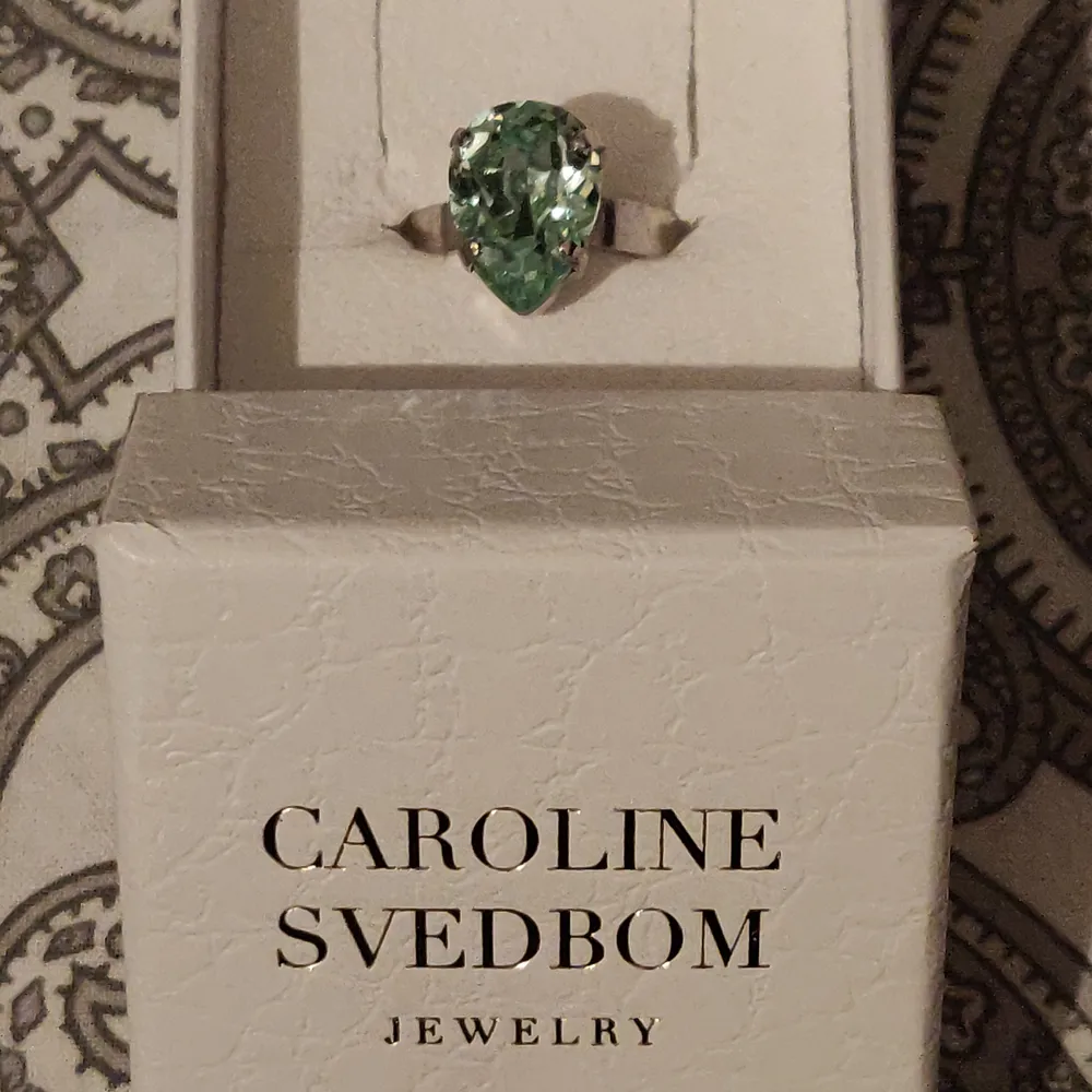 Mini Drop Ring Rhodium Peridot Delite 495 kr  Mini Drop Ring Rhodium Peridot Delite från Caroline Svedbom. En öppen ring med en vacker droppformad Swarovskisten i en kungligt, ljus grön färg.   Mått: 13 x 10 mm Storlek: One size, justerbar Material: Rhodiumpläterad mässing & Swarovisk-kristall. Accessoarer.
