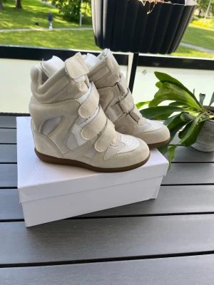 Isabel Marant vita wedge sneakers - Snygga vita wedge sneakers från Isabel Marant med beige mockadetaljer och kardborreband. Skorna har en chunky sula i gummi och cool high-top design som ger extra höjd. Perfekta för dig som vill ha en trendig och unik look.