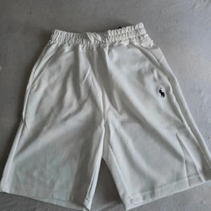 Vita shorts från Polo Ralph Lauren - Säljer ett par klassiska vita shorts från Polo Ralph Lauren med elastisk midja och sidofickor. Shortsen har den ikoniska loggan broderad på vänster ben och är tillverkade i mjuk bomull för en skön känsla.