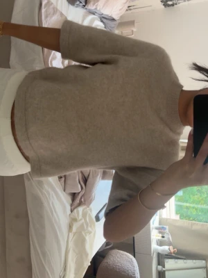 Beige stickad tröja med kort ärm - Beige stickad tröja med korta ärmar och rund hals. Tröjan har en rak, avslappnad passform och är gjord i ett mjukt, tunt material som känns skönt mot huden. Enkel och stilren design som är lätt att matcha.
