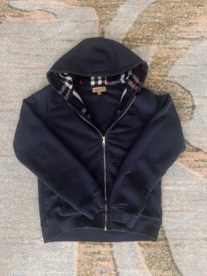 Mörkblå hoodie från Burberry - Snygg mörkblå hoodie från Burberry med dragkedja och klassiskt rutigt foder i huvan. Hoodien har ribbade muddar, två fickor framtill och är tillverkad i mjukt bomullsmaterial. Perfekt för en chill och stilren look. Tyvärr finns inte snöre vilket reducerar priset.