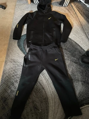 Svart Nike loungewear set med huva - Säljer ett svart loungewear set från Nike med gul logga. Setet består av en hoodie med dragkedja och fickor samt matchande byxor med resår i midjan och sidofickor. Både tröjan och byxorna har gula Nike-detaljer. Perfekt för chill dagar hemma.