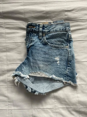 Blå jeansshorts med slitningar - Säljer ett par blå jeansshorts med råa, fransiga kanter och slitna detaljer framtill. Klassisk femficksmodell med dragkedja och knapp. Perfekta för en avslappnad och trendig sommarlook.