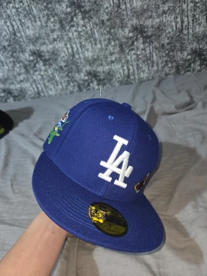 Blå LA New Era keps med brodyr - Säljer en blå New Era 59FIFTY keps med LA-logga i vitt framtill. Kepsen har platt skärm och är dekorerad med broderade fjärilar och figurer på sidan. Perfekt för dig som vill sticka ut med en unik accessoar.