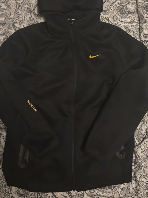 Nike nocta tracksuit - Snygg svart hoodie från Nike NOCTA-kollektionen med gul Nike-logga på bröstet och NOCTA-tryck på ärmen. Dragkedja framtill, två fickor med dragkedja och huva. Materialet är mjukt och följsamt, perfekt för en streetwear-look.