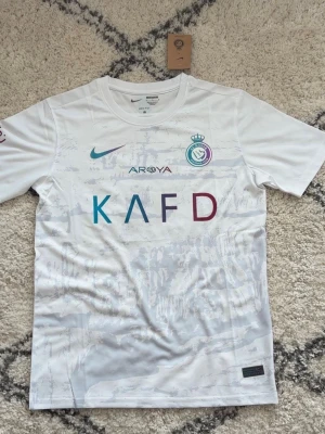 Vit Nike Al Nassr matchtröja - Snygg vit matchtröja från Nike för Al Nassr FC med klubbmärke och sponsortryck i flera färger på bröstet. Tröjan har rund hals, korta ärmar och är tillverkad i ett lätt, andningsbart funktionsmaterial som passar perfekt till träning eller match.