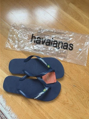 Mörkblå flip-flops från Havaianas - Snygga mörkblå flip-flops från Havaianas med klassisk design och bekväm gummisula. Remmarna har Havaianas-logga och en liten brasiliansk flagga. Perfekta för sommaren och strandhäng! Storlek 43/44