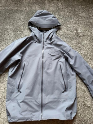 Arcteryx Beta SL Jacka - Observera att jackan kan se grå ut på bilderna på grund av ljus och kamera, men den verkliga färgen är en snygg blå/ljusblå nyans.  Säljer min Arc’teryx Beta SL, en lätt och smidig skaljacka av hög kvalitet. Perfekt för både vardag, vandring och friluftsliv. Jackan är vind- och vattentät med ett tekniskt material som håller dig torr även i tuffare väder.  Passar även perfekt under sommaren och varmare månader, då den är lätt, andas bra och funkar utmärkt vid regniga eller blåsiga dagar.