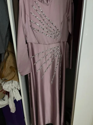 Ljuslila långklänning med strassdetaljer - Elegant ljuslila långklänning med långa ärmar och iögonfallande strassdetaljer i solfjädersmönster över bröstet och höften. Klänningen har en mjuk, glansig finish och markerad midja för en snygg siluett. Perfekt för dig som vill sticka ut.