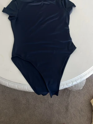Mörkblå kortärmad body - Snygg mörkblå body med korta ärmar och rund halsringning. Tillverkad i ett glansigt, stretchigt material som sitter tight mot kroppen. Perfekt att styla med jeans eller kjol för en trendig look.