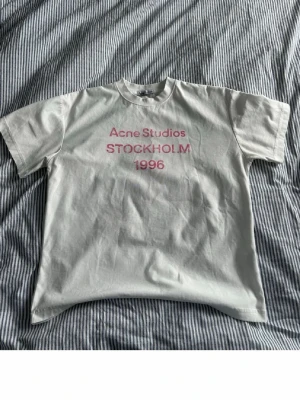 Vit Acne Studios t-shirt Stockholm - Vit t-shirt från Acne Studios med rosa text 'Acne Studios STOCKHOLM 1996' på bröstet. Klassisk passform med korta ärmar och rund hals. Perfekt basic med en clean look och snygg branding framtill.
