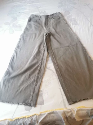 Gråa wide leg jeans med råa kanter - Säljer ett par gråa wide leg jeans med hög midja och råa, fransiga kanter nertill. Jeansen har klassiska fickor både fram och bak och är tillverkade i ett mjukt denimtyg. Perfekta för en avslappnad och trendig look.