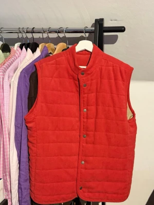 Röd quiltad manchesterväst från Morris  - Stilren röd väst i manchester med quiltad design och tryckknappar framtill. Hög krage och normal passform gör den enkel att bära över en hoodie eller skjorta. Perfekt för dig som vill ha en färgklick i garderoben.