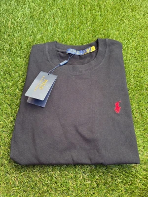 Svart t-shirt från Polo Ralph Lauren - Klassisk svart t-shirt från Polo Ralph Lauren med röd broderad logga på bröstet. T-shirten har rund hals och korta ärmar, perfekt för en clean och stilren look. Tillverkad i mjuk bomull för skön komfort. Size ”M”