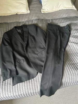 Svart kostym med kavaj och byxor - Klassisk svart kostym med matchande kavaj och byxor. Kavajen har smala slag, två framfickor och knappar vid ärmslut. Perfekt för dig som vill ha en stilren och tidlös look. Passar till många olika tillfällen. Kavaj storlek 54 byxa 34/32
