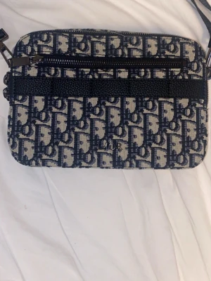 D¡or axelväska i blått monogram - Snygg axelväska från D¡or med klassiskt blått och beige monogrammönster. Väskan har en rektangulär form, silverfärgad logga framtill och dragkedja upptill. Tillverkad i canvas med justerbar axelrem. Perfekt för dig som gillar lyxiga detaljer och trendig design.