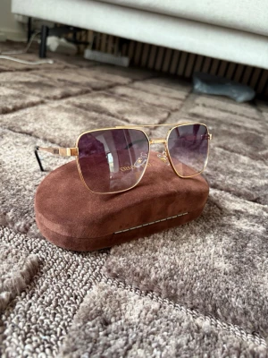  solglasögon guld - Snygga solglasögon från  med fyrkantiga guldiga metallbågar och lila tonade glas. Kommer med ett brunt fodral i mockaimitation. Perfekta för att lyfta din stil och skydda ögonen mot solen. Obs fodralet ingår inte utan är för att lyfta glasögonen