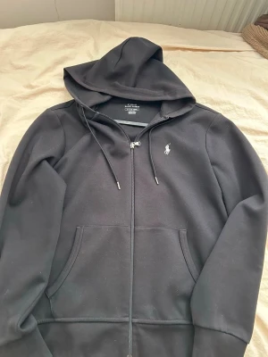 Svart hoodie från Polo Ralph Lauren - Snygg svart hoodie från Polo Ralph Lauren med dragkedja framtill och klassisk vit logga på bröstet. Hoodien har huva med dragsko och två stora fickor framtill. Tillverkad i mjukt bomullsmaterial som känns skönt mot huden.
