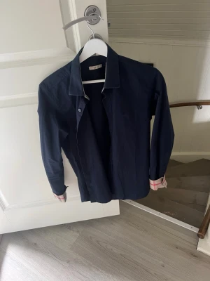 Mörkblå skjorta med rutiga detaljer - Stilren mörkblå skjorta med långa ärmar och klassisk krage. Insidan av manschetterna har snygga rutiga detaljer i beige, rött och vitt. Perfekt för dig som gillar en enkel men ändå unik look. storlek M men skulle säga att den passar bättre för S