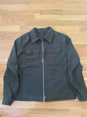 Olivgrön overshirt från Massimo Dutti - Stilren olivgrön overshirt från Massimo Dutti med två bröstfickor med lock och knapp, samt dragkedja framtill. Tillverkad i ett tåligt bomullsmaterial och har klassisk krage. Perfekt lager-på-lager-plagg för en avslappnad look.