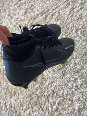 Nike Air Zoom svarta fotbollsskor - Säljer ett par svarta Nike Air Zoom fotbollsskor med högt skaft och snörning. Skorna har en modern design med diskret Nike-logga på sidan och mönstrad ovandel. Yttersulan har dobbar för bra grepp på planen. Perfekta för dig som vill ha stilrena och funktionella skor.
